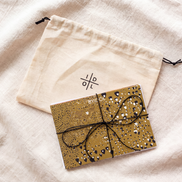 A gold glitter notebook tied with a black elastic string, placed next to a cream-colored cotton drawstring pouch with a black symbol logo, both resting on a textured beige linen fabric background.
