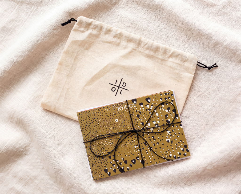A gold glitter notebook tied with a black elastic string, placed next to a cream-colored cotton drawstring pouch with a black symbol logo, both resting on a textured beige linen fabric background.