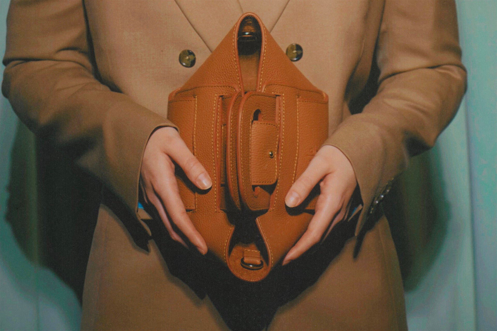 Person wearing a brown coat holding a brown leather bag against a light blue background